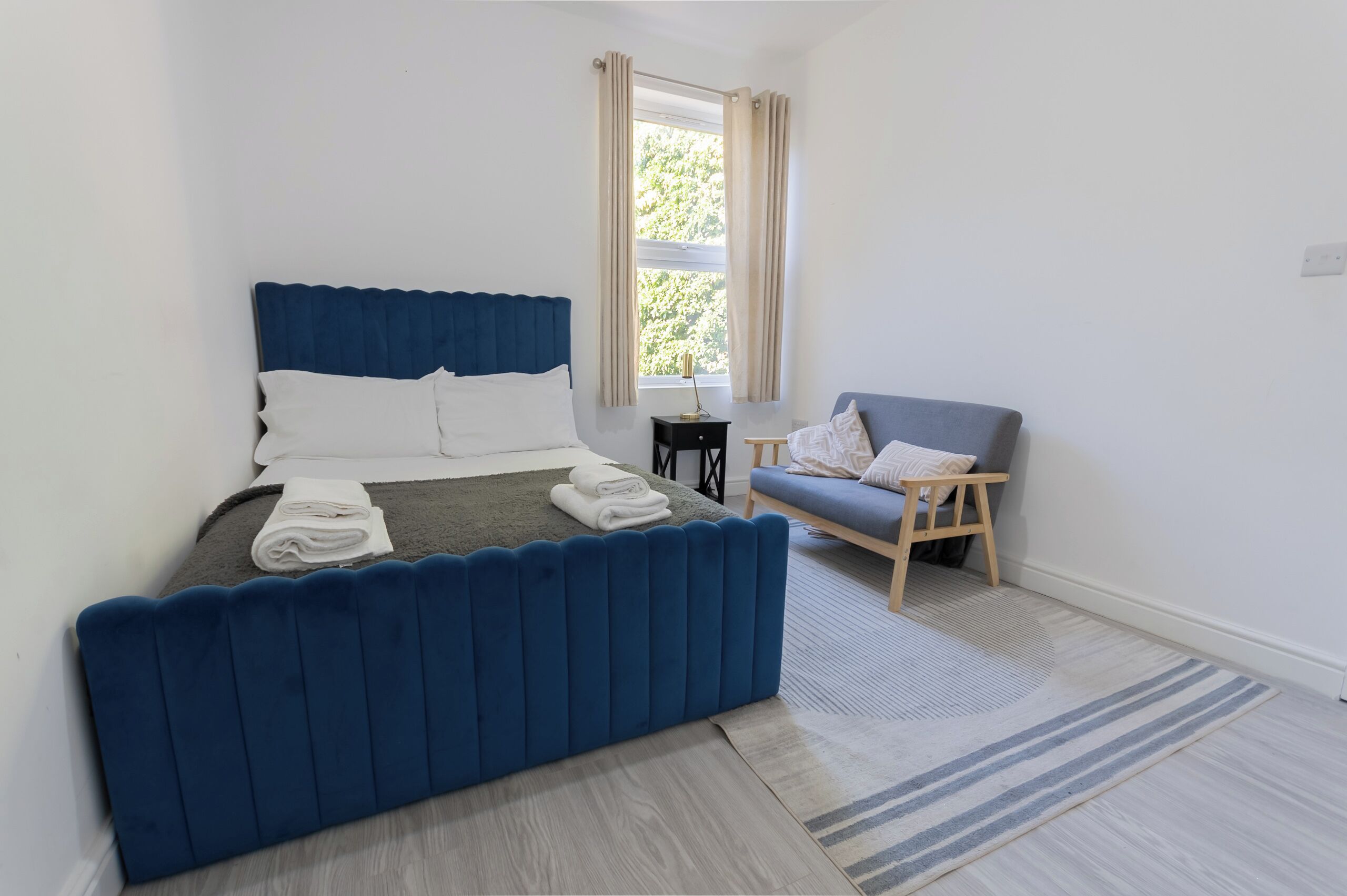 Une chambre à coucher avec un lit recouvert d’un tissu bleu, une petite table de chevet en bois, une chaise et une fenêtre avec des rideaux.