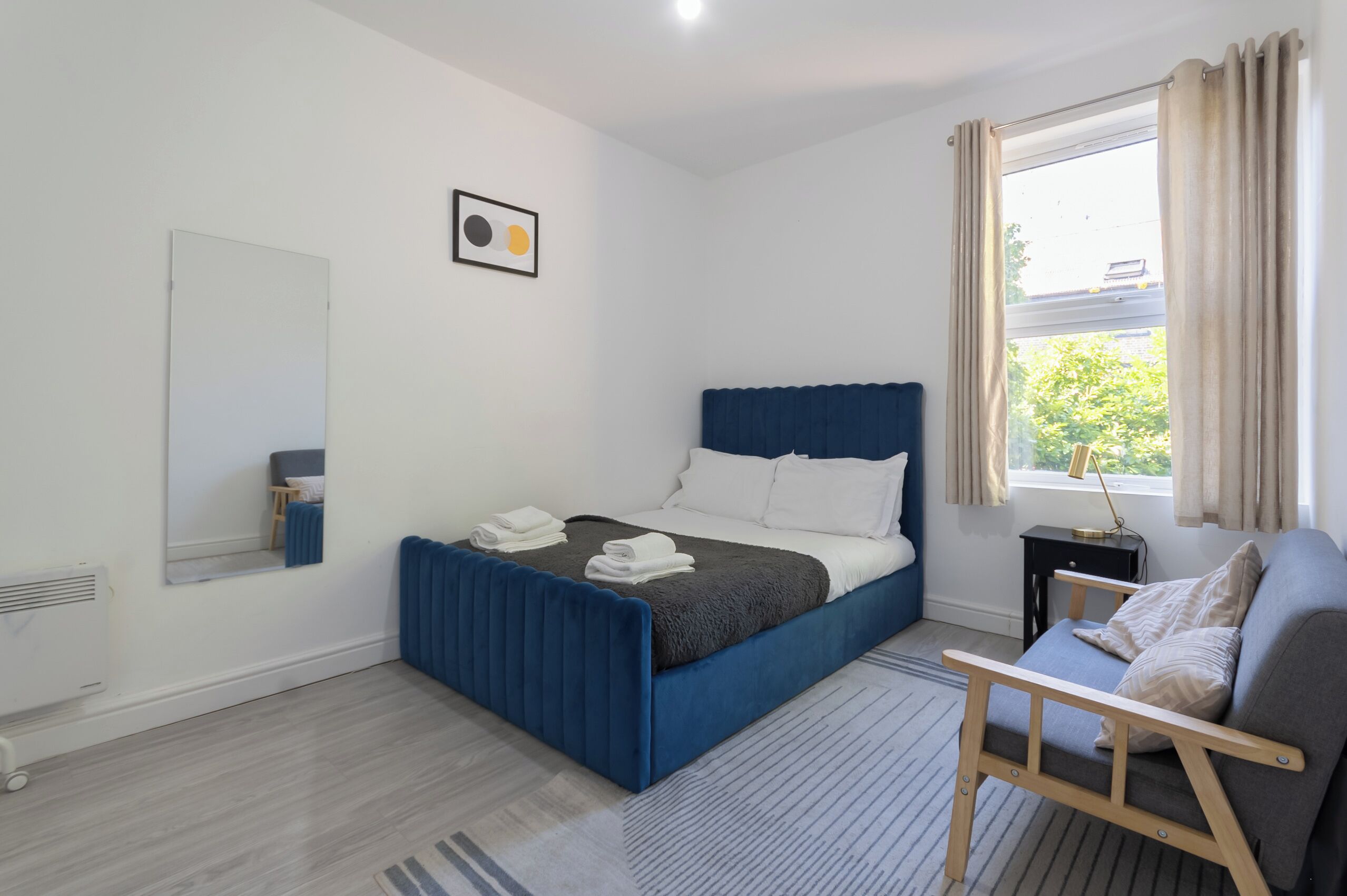 Een slaapkamer met een blauw bed, een nachtkastje, een stoel, een raam met gordijnen, en een spiegel aan de muur.