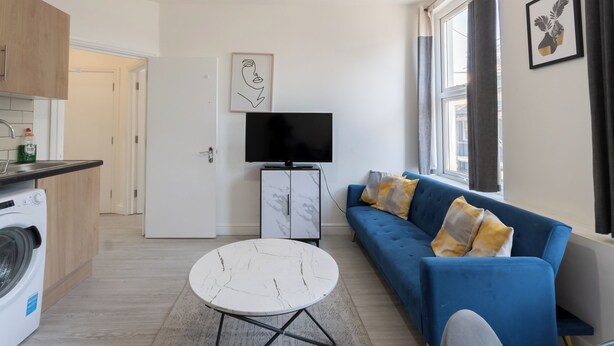 A modern living room with a blue sofa, a white marble coffee table, a flat-screen TV, and a kitchen area with wooden cabinets and a washing machine.