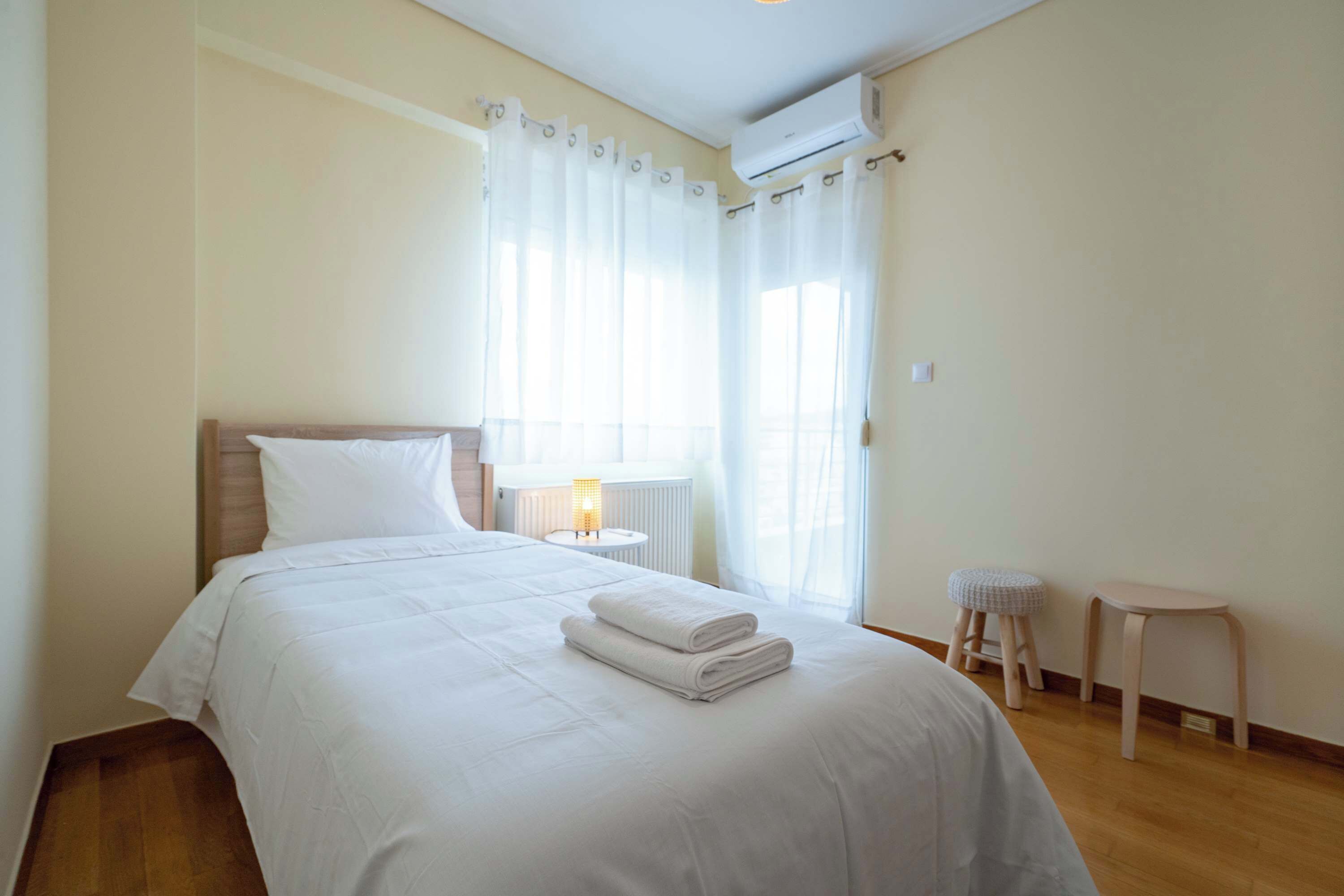 Una cama bien hecha con sábanas y toallas blancas, una mesita de noche con lámpara, una ventana con cortinas translúcidas y un radiador.