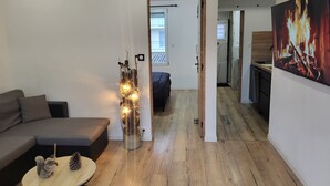 A modern living room with a grey sofa, a small round table with a pinecone, and a decorative light fixture. A hallway leads to a kitchen area with a large painting of fire.