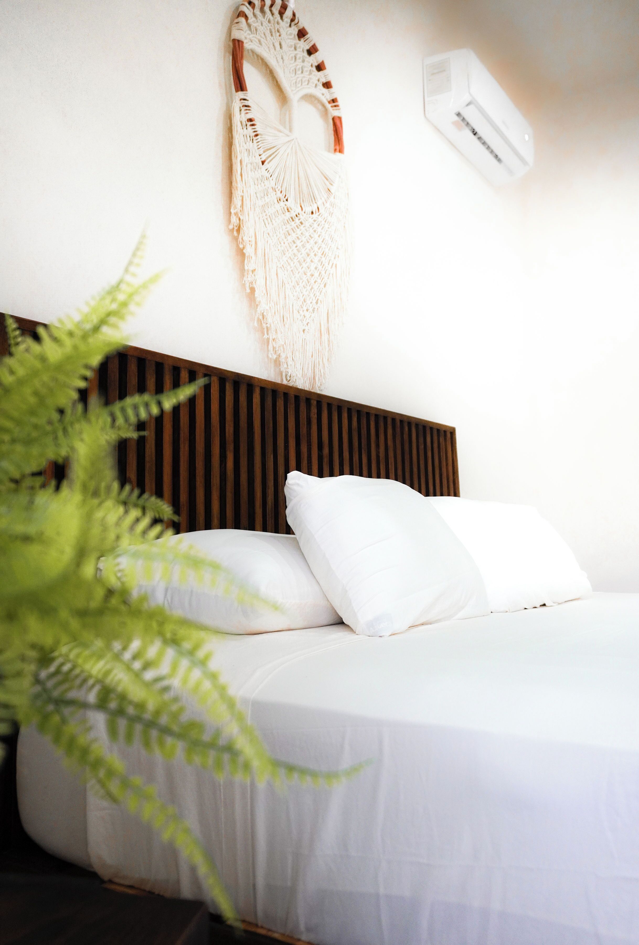 Una cama bien hecha con sábanas y almohadas blancas, un tapiz de macramé y un aire acondicionado de pared.