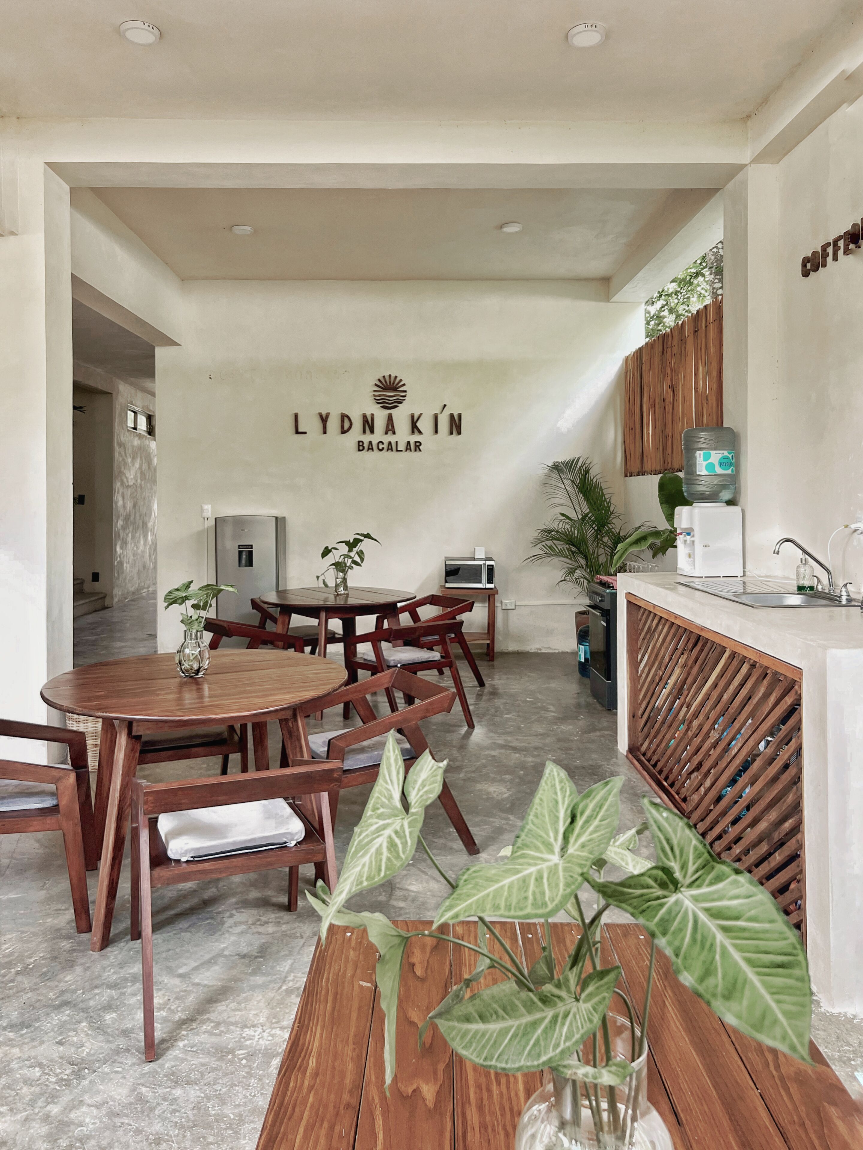 A modern dining area with wooden furniture, a water dispenser, and a sign reading "LYDNAKIN BACALAR".