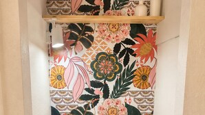 Escrivaninha no canto com papel de parede floral, cadeira rosa e prateleira com objetos decorativos.