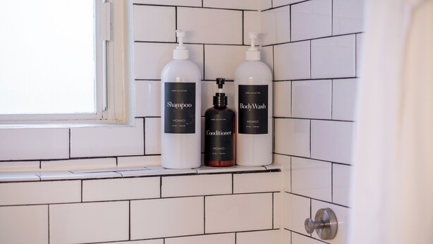 Un bagno con pareti piastrellate in bianco e una finestra. Si vedono tre prodotti Moikko: shampoo, balsamo e detergente corpo.