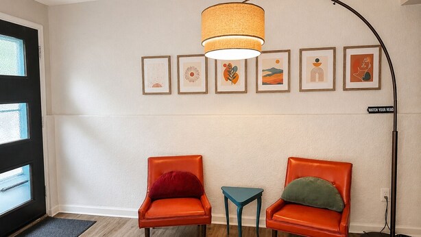 Quarto com duas cadeiras laranja, uma mesinha lateral azul-esverdeada, um abajur e seis quadros na parede.