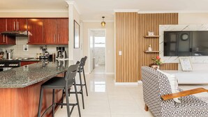 Eine moderne Küche mit einer Granit Arbeitsplatte, Holzschränken und einem eingebauten Herd. Es gibt eine Essecke mit Barhocker und eine Couch mit gemustertem Stoffbezug. Ein Flachbildfernseher ist über der Couch an der Wand befestigt.