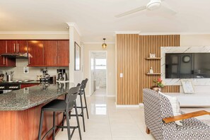 A modern kitchen with a granite countertop, wooden cabinets, and a built-in stove. There is a dining area with bar stools and a sofa with a patterned upholstery. A flat-screen TV is mounted on the wall above the sofa. - Bella View of Table Mountain to make your stay even more magical (Cape Town)