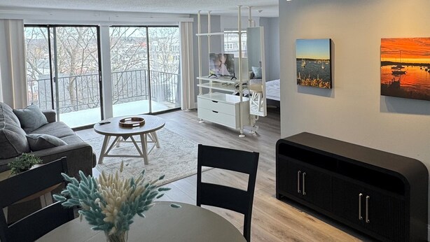 Een moderne woonkamer met een groot raam, een eethoek met een ronde tafel en een zwarte entertainmentkast tegen de muur.
