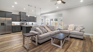 Eine moderne Küche mit dunklen Schränken und Edelstahlgeräten, ein graues Ecksofa mit gemusterten Kissen und ein Holztisch.