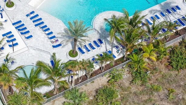 Luftaufnahme eines Resorts mit einem zentral gelegenen Swimmingpool, umgeben von Liegestühlen und Palmen.