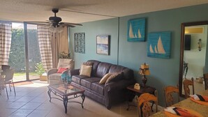Ein Wohnzimmer mit einer Ledersofa, einem Glastisch und einem Esstisch mit Stühlen. Vorhanden sind Vorhänge, ein Deckenventilator und Kunstwerke an den Wänden.