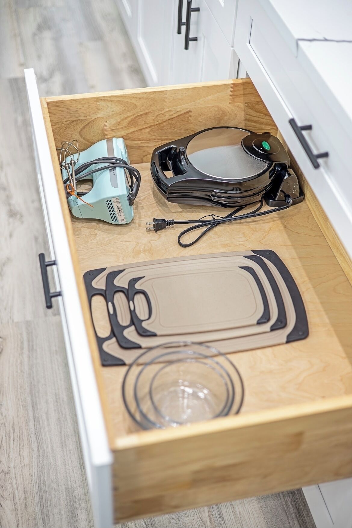 Un cajón con una tabla de cortar de madera, un vaso y un electrodoméstico negro.
