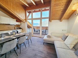 A modern kitchen with wooden beams, a dining area with a table and chairs, and a sofa with a view of the mountains.