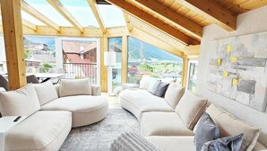 Een ruime kamer met een hoge plafond en houten balken, met een grote hoekbank en uitzicht op een bergachtig landschap.