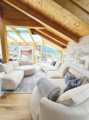 A spacious room with a high ceiling and wooden beams, featuring a large sectional sofa and a view of a mountainous landscape.