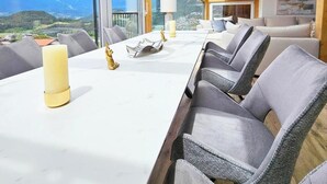 Een kamer met hoge plafonds en een houten balkenstructuur, een lange eettafel met stoelen en uitzicht op bergen door grote ramen.