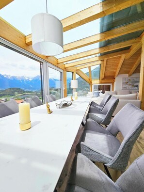 A high-ceilinged room with a wooden beam structure, a long dining table with chairs, and a view of mountains through large windows.