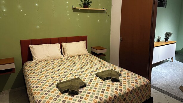 A bedroom with a bed, two pillows, a bedside table, a nightstand, a dresser, and two potted plants. - Seu cantinho na Gamboa (Cabo Frio)