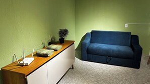 A room with a wooden desk, a blue sofa, a green wall, and a potted plant. - Seu cantinho na Gamboa (Cabo Frio)