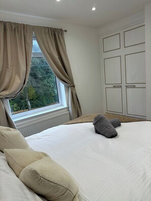 Un dormitorio con una cama grande, cortinas color beige, una ventana con vistas a árboles y un armario blanco.