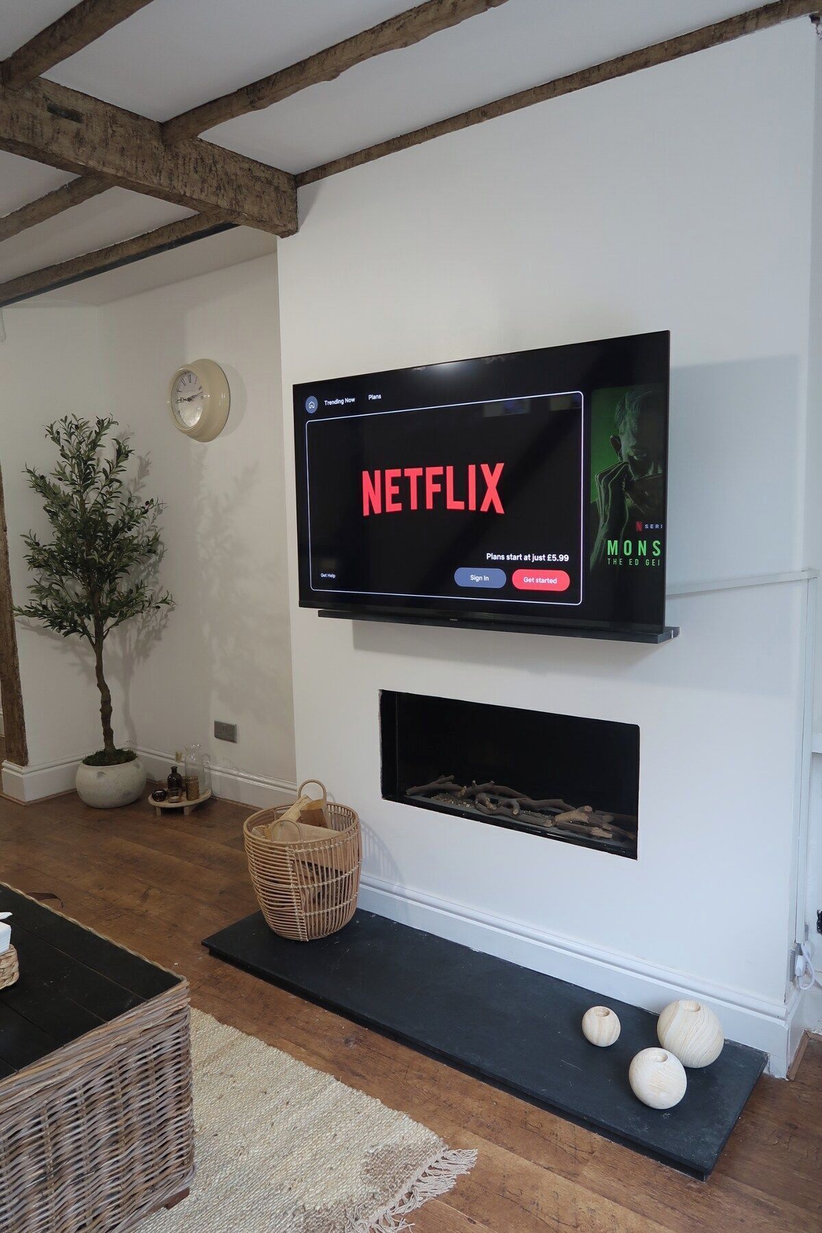 A living room with a mounted TV displaying a Netflix interface, a fireplace below, a potted plant, and a wicker basket.