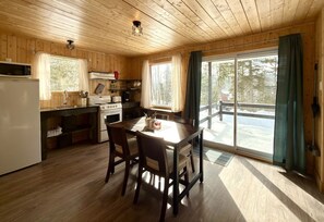 A wooden cabin interior with a kitchen, dining area, and large sliding glass doors to a deck. - Mini Chalet at Lake Pohénégamook, a Peaceful Haven! #1 (Pohénégamook)