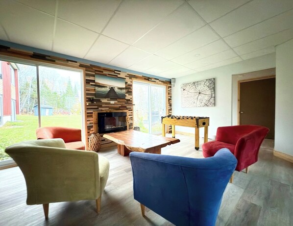 A modern living room with a fireplace, a foosball table, and a large window offering a view of the outdoors.