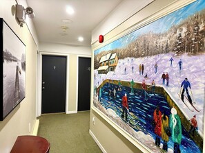 A hallway with a large painting of skiers on a snowy slope, two doors, and a red object on the wall. - Grand Havre – Perfect for Large Gatherings (Pohénégamook)