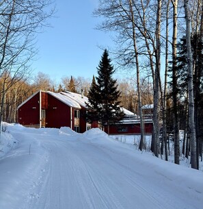 A red barn with a snow-covered roof, surrounded by trees and a snow-covered path.