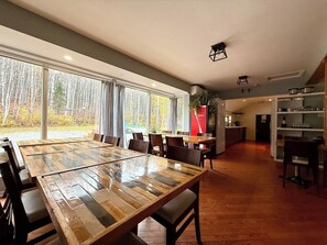 Un vaste espace à manger avec une grande table et des chaises en bois, une cuisine en arrière-plan et une vue sur les arbres à travers de grandes fenêtres.