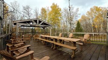 An outdoor wooden deck with picnic tables and chairs, a covered seating area, and a view of trees.