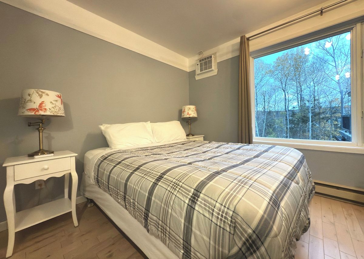 Een slaapkamer met een bed, twee nachtkastjes met lampen, een raam met uitzicht op bomen, en een wandairconditioner.