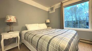 Een slaapkamer met een bed, twee nachtkastjes met lampen, een raam met uitzicht op bomen, en een wandairconditioner.