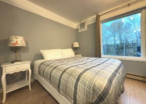 Ein Schlafzimmer mit einem Bett, zwei Nachttischen mit Lampen, einem Fenster mit Blick auf Bäume und einer wandmontierten Klimaanlage.