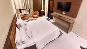 Quarto de hotel com uma cama, uma televisão na parede, uma escrivaninha e duas cadeiras.