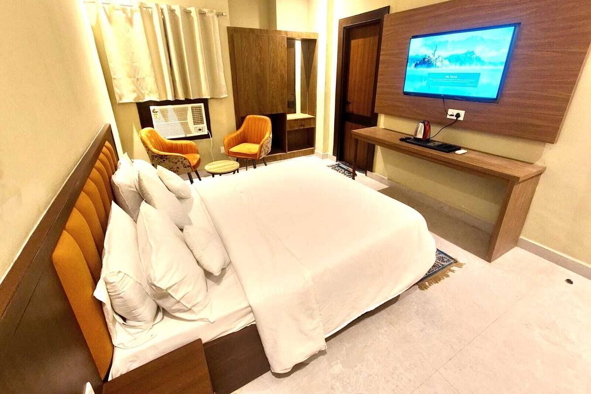 A hotel room with a bed, two armchairs, a small table, a TV on the wall, and a window with curtains.