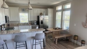 Una cocina moderna con armarios blancos, encimera de granito, una isla central con taburetes altos, una mesa de comedor de madera y un amplio ventanal que proporciona luz natural.