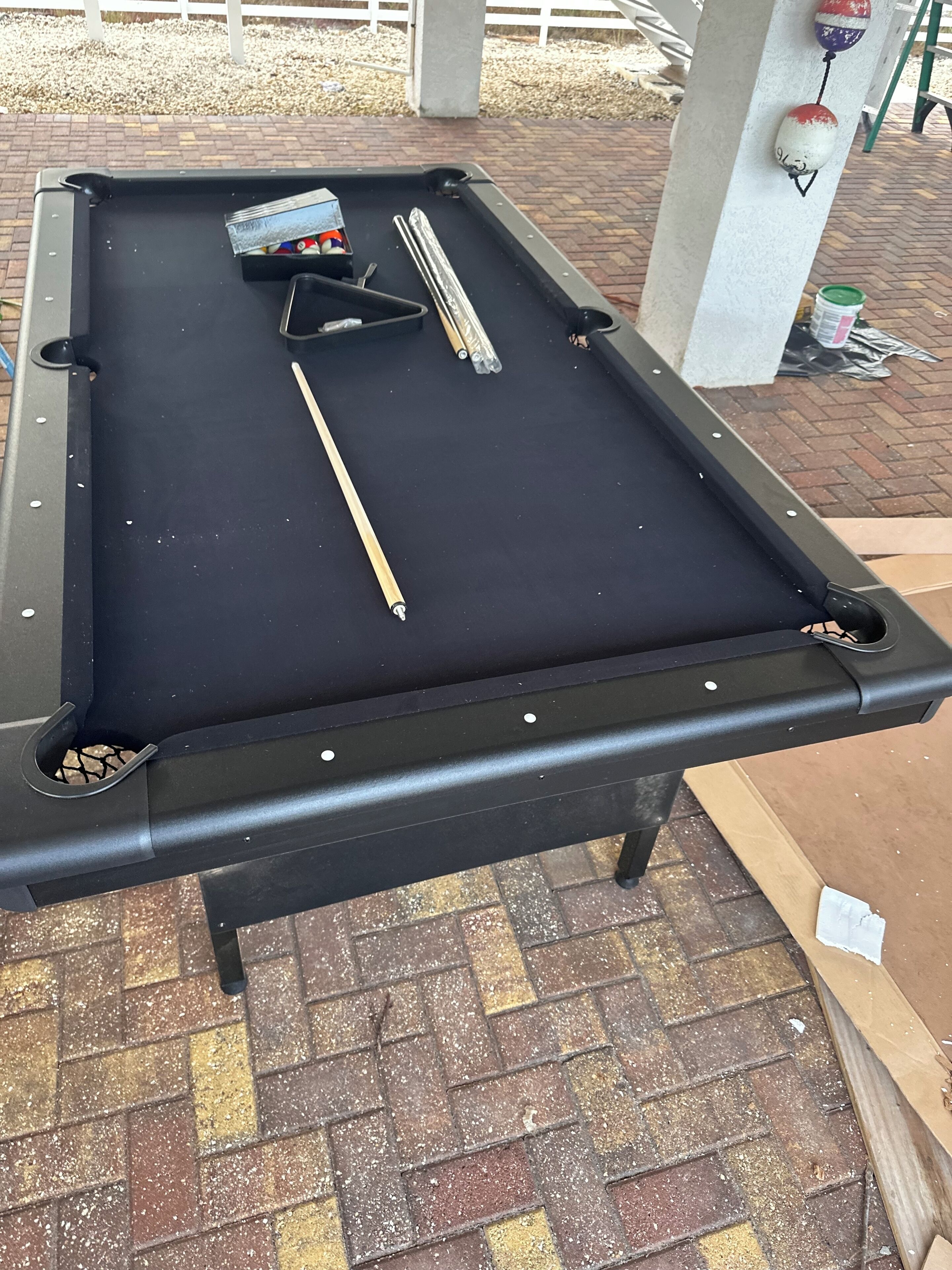 A pool table with billiard balls and cues, surrounded by a brick floor and a white fence in the background.