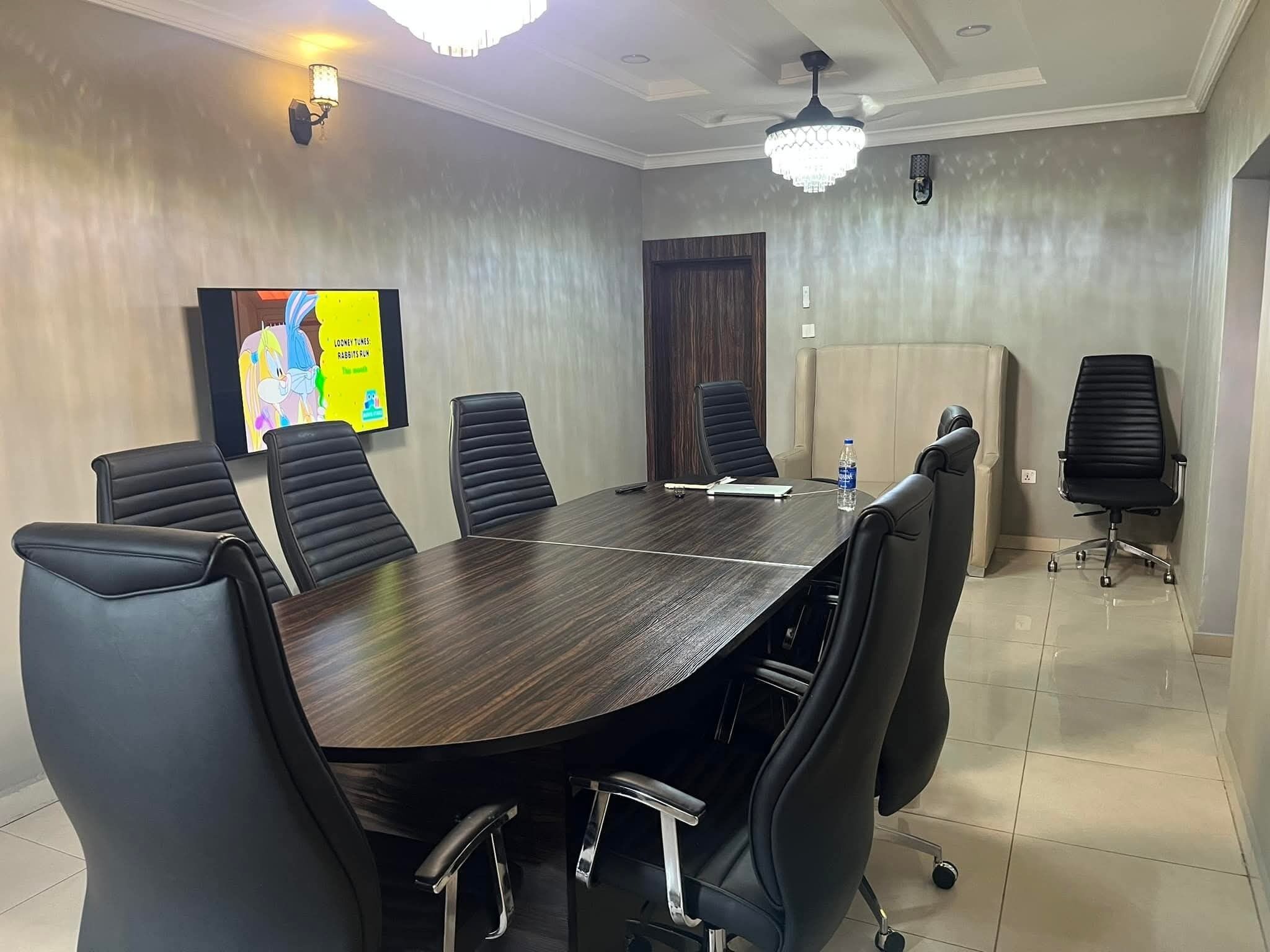 Une salle de conférence avec une grande table en bois foncé, des fauteuils en cuir noir, un grand écran et un lustre.