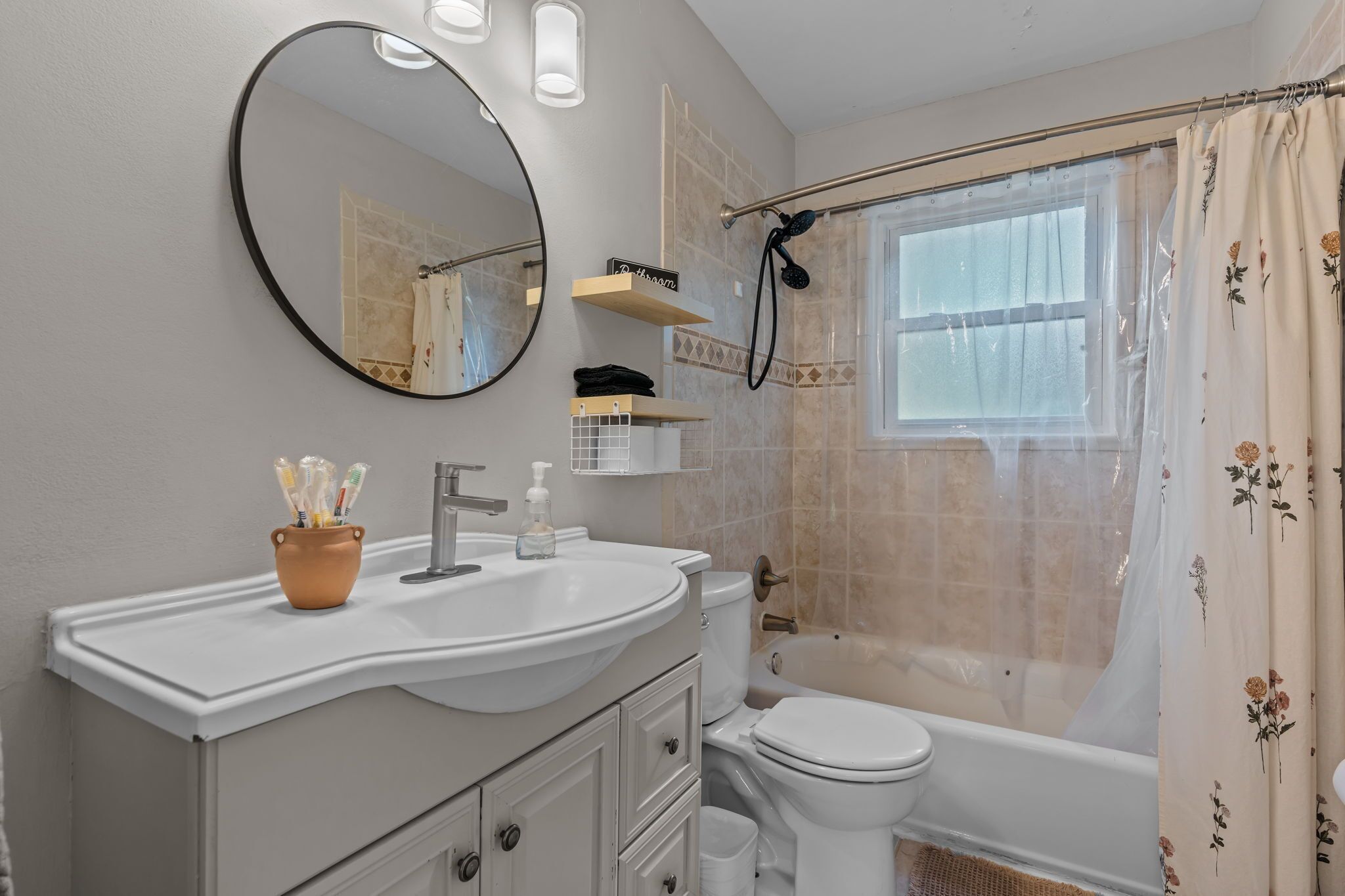 A bathroom with a white sink, a toilet, a bathtub, and a shower. There are shelves above the sink and a shower curtain with floral patterns.