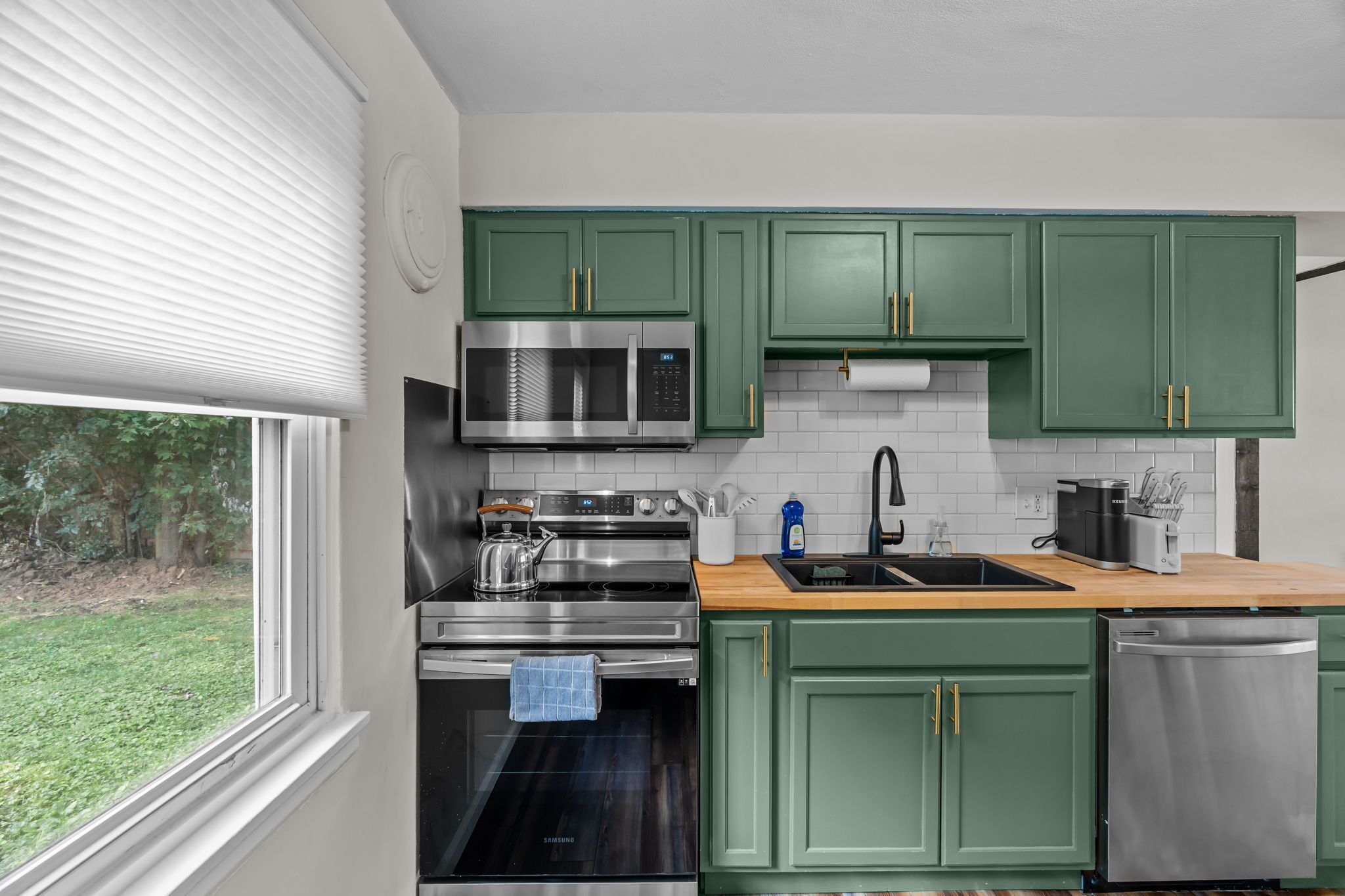 Una cocina moderna con gabinetes verdes, electrodomésticos de acero inoxidable y una ventana con vistas a un jardín.