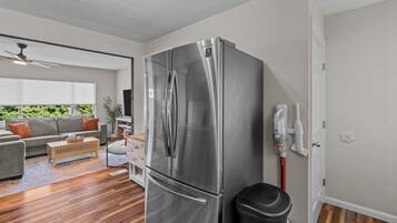 A modern kitchen with a stainless steel refrigerator, wooden flooring, and a grey sofa in the adjacent living area.