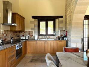 A kitchen with wooden cabinets, a stainless steel range hood, a marble countertop, and a window with a view of greenery. - Luxury Crete Villa with Infinity Pool, Hot Tub & White Mountain Views (Douliana)