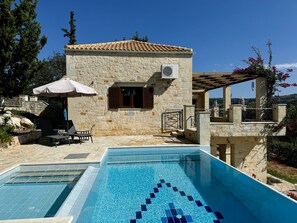 Un edificio de piedra con techo de tejas, piscina con diseño de mosaico azul, mobiliario exterior y una pérgola con flores rojas.