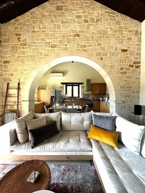 A living room with a stone wall, an arched doorway leading to a kitchen, and a sofa with cushions. - Luxury Crete Villa with Infinity Pool, Hot Tub & White Mountain Views (Douliana)