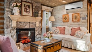 A cozy living room with a stone fireplace, a white sofa with floral cushions, a vintage trunk coffee table, and a mounted air conditioner. - Boulder Hot Tub Buffalo River Fairy Tale  Cottage (Jasper)