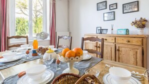 Uma sala de jantar com uma mesa de madeira posta para o café da manhã, com pratos, tigelas e uma fruteira. Cadeiras estão dispostas ao redor da mesa, há uma janela com cortinas e um armário com quadros.