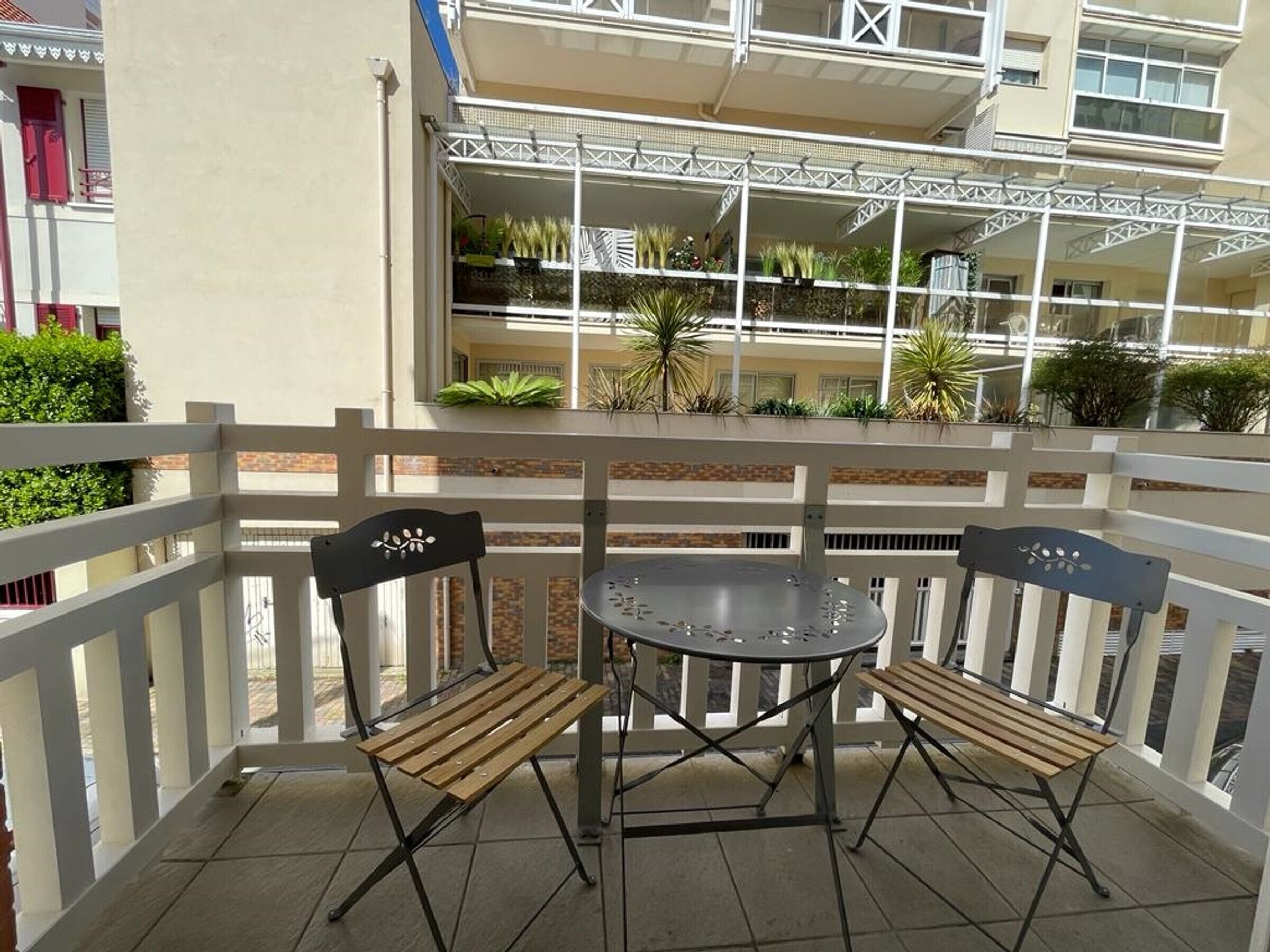 A balcony with a table and two chairs, a white railing, and a building with multiple balconies and plants in the background.