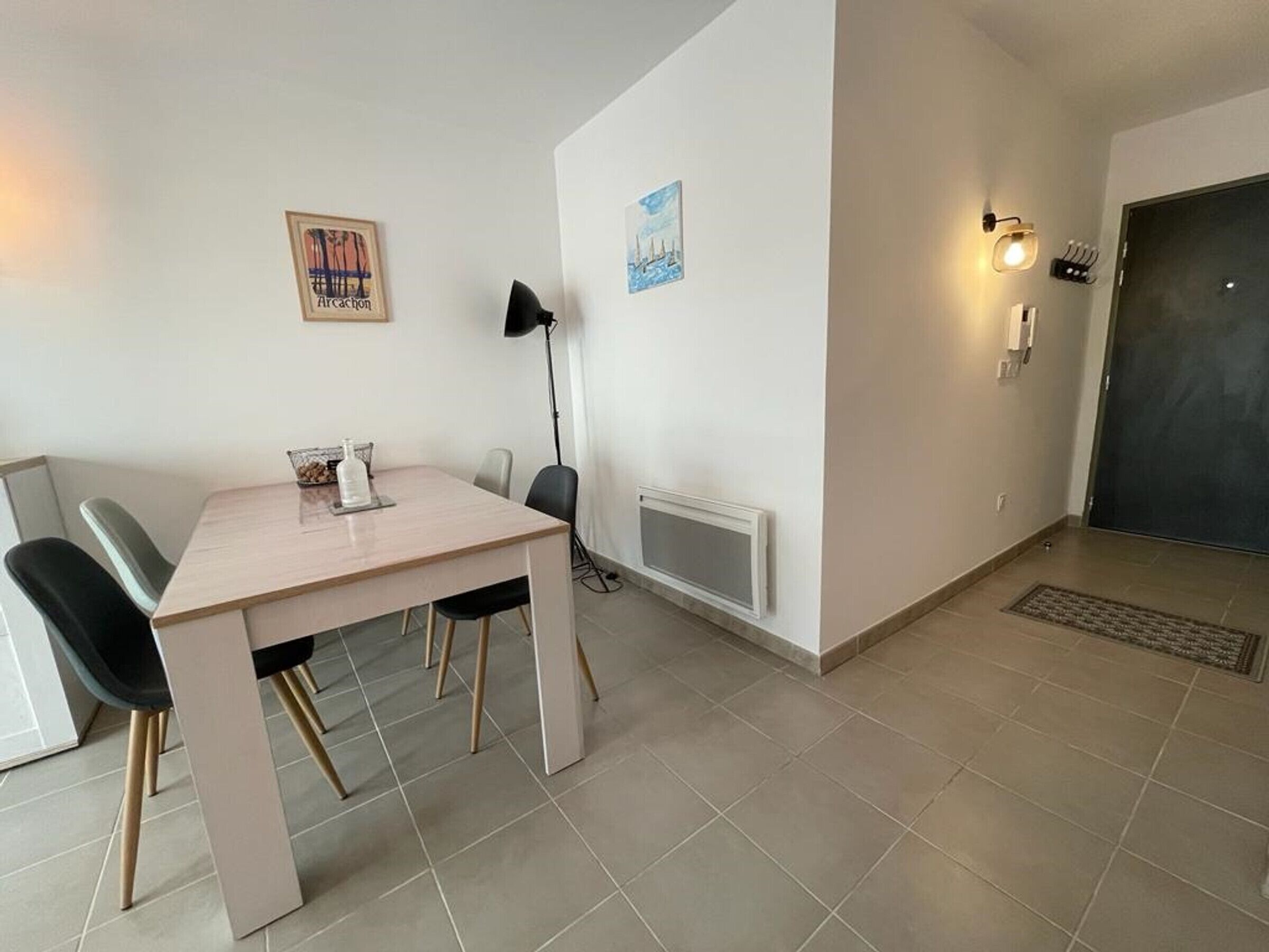A modern, minimalist dining area with a light-colored table and chairs, a floor lamp, and two framed pictures on the walls.